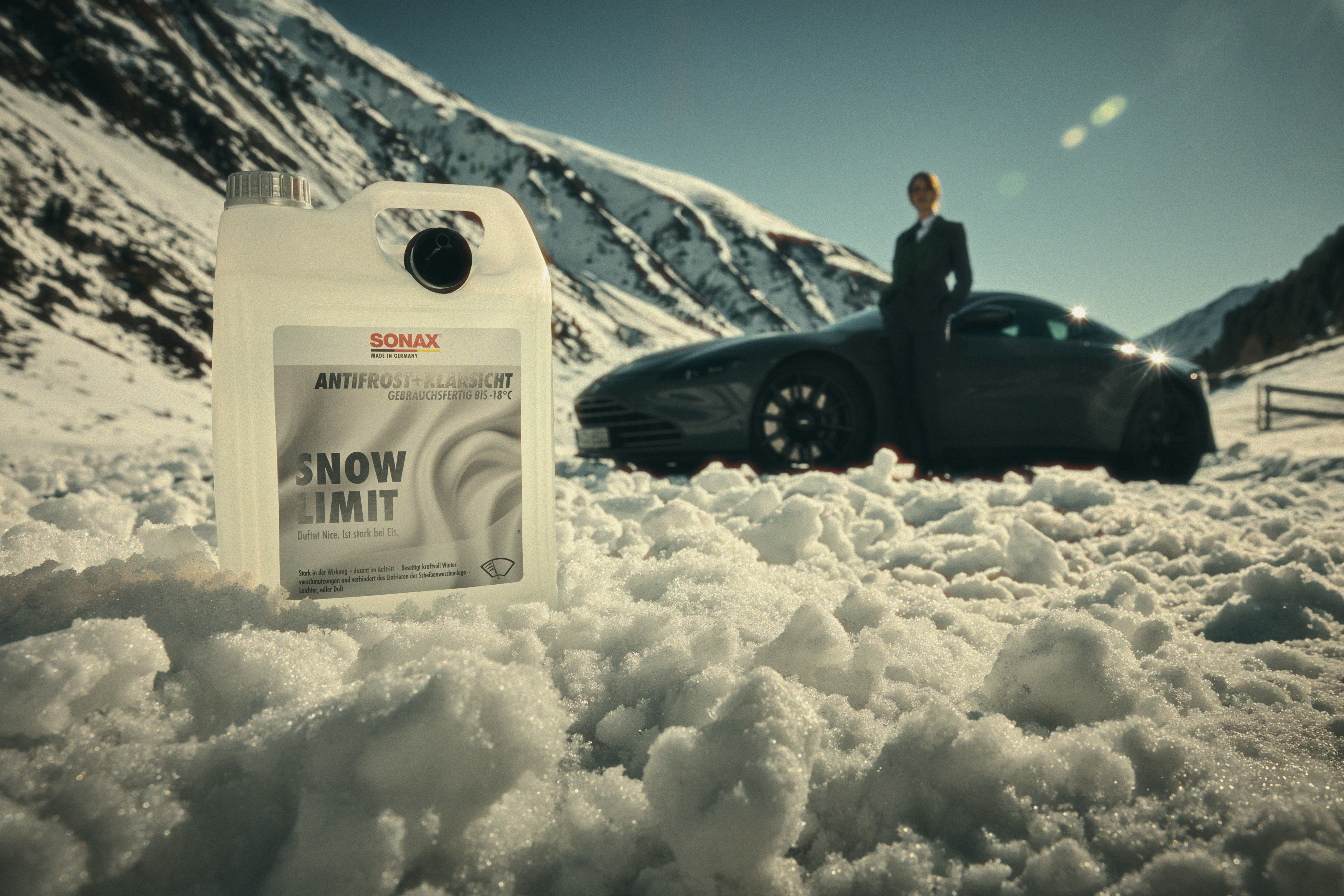 Eine Schneelandschaft mit Berg im Hintergrund. Im Vordergrund ein weißer 5 Liter Kanister mit Aufdruck 'Snow Limit' in schwarz. Unscharf im Hintergrund ein Schwarzes Auto, vor dem die dunkle Silhouette einer Person zu sehen ist.