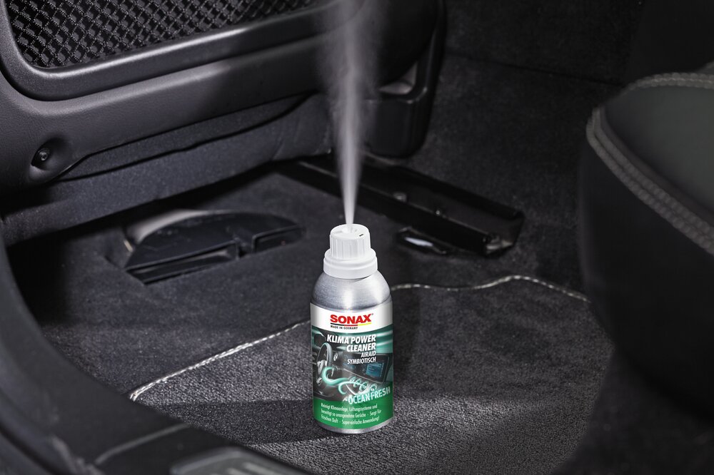 Ein SONAX KlimaPowerCleaner AirAid steht im Fußraum und sprüht.