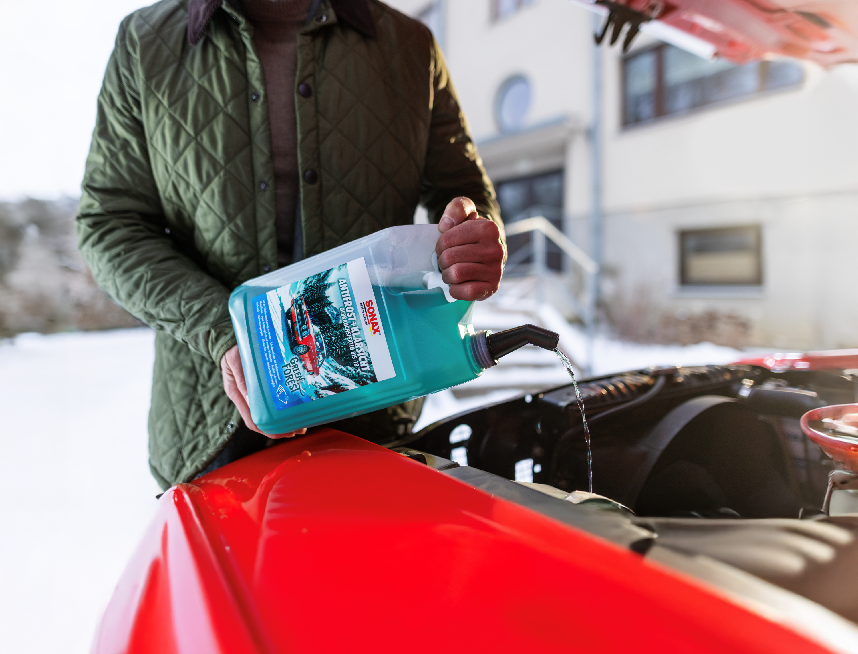 Der SONAX Antifrost+Klarsicht in der Duftrichtung Green Forest steht in einer verschneiten Wald mit einem roten Oldtimer im Hintergrund.