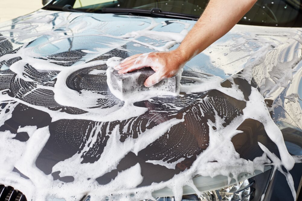 Ein graues Auto ist mit Autoshampoo eingeschäumt und eine Person wäscht das Auto mit einem Schwamm