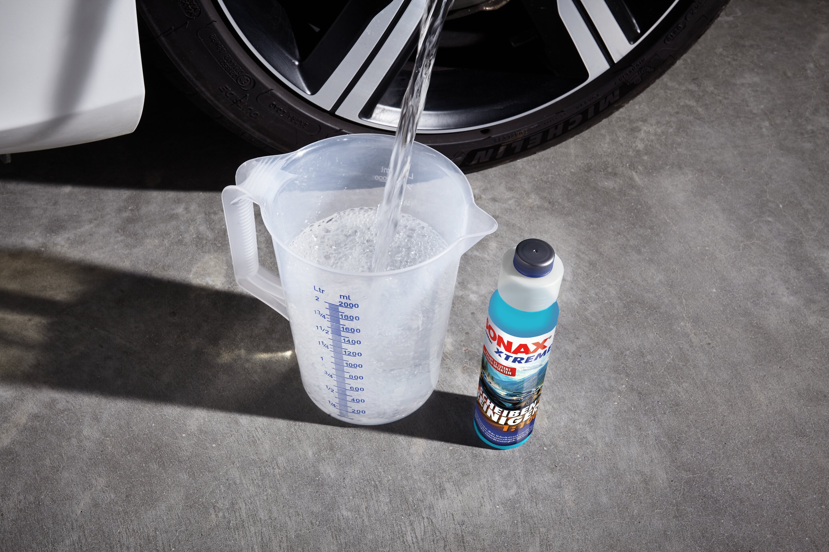Ein Messbecher und der XTREME Schei­ben­Rei­niger 1:100 stehen vor einem Autoreifen. Ein Wasserstrahl befüllt den Messbecher.