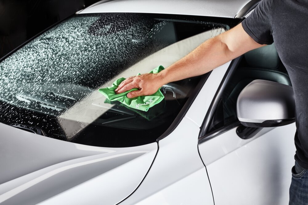 Die Frontscheibe eines Autos wurde mit ScheibenKlar besprüht und jemand wischt die Scheibe mit einem Mikrofasertuch nach.