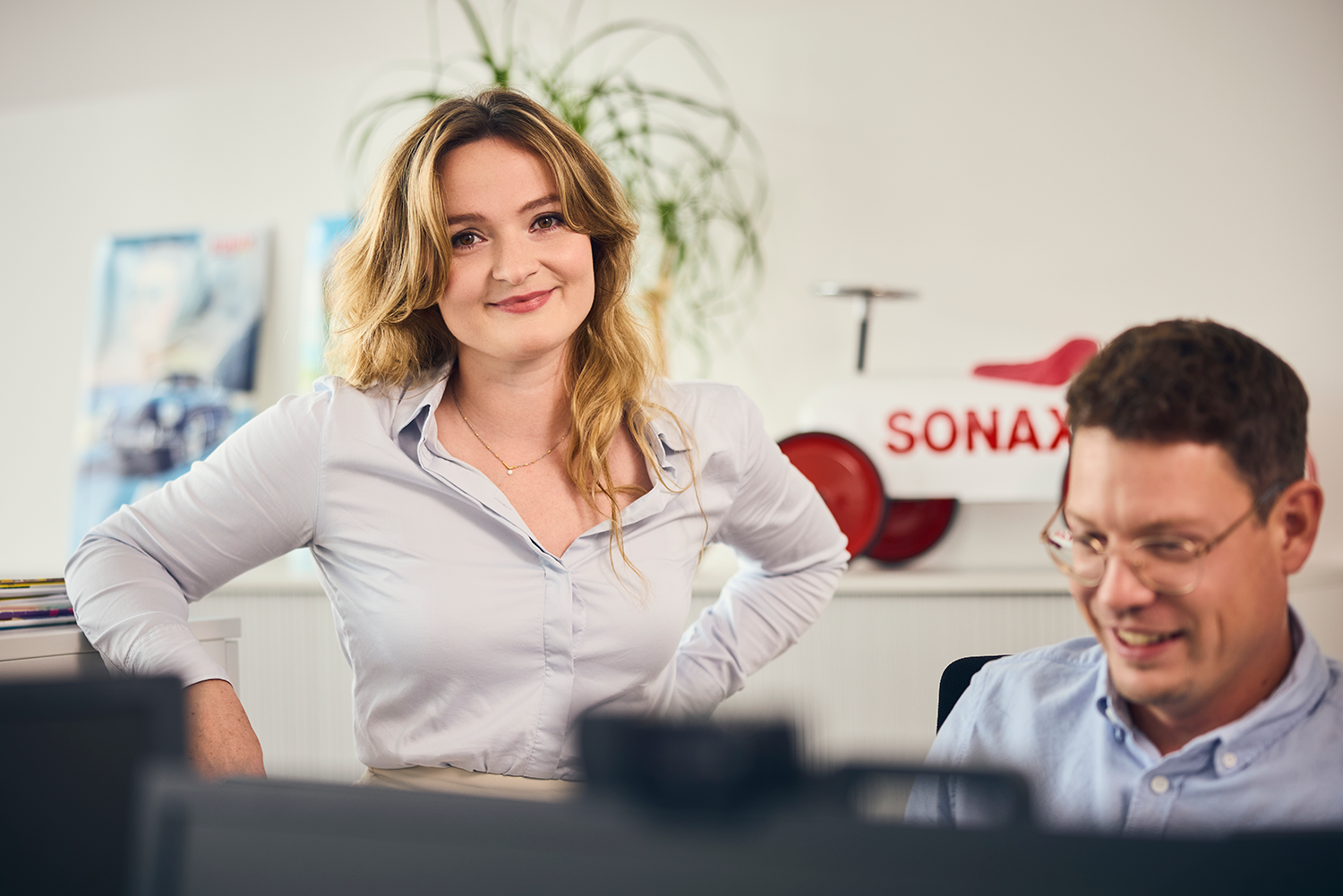 Eine Frau steht neben einem Mann der am Computer arbeitet und lächelt in die Kamera
