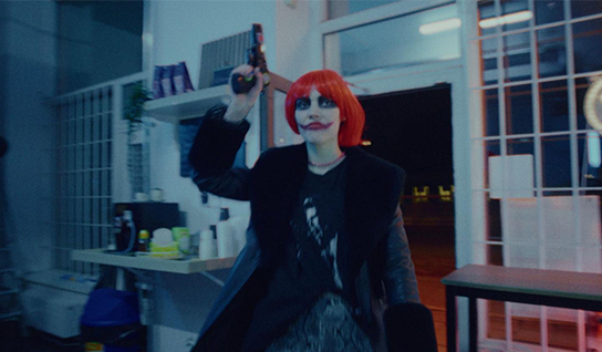 A woman with a red wig and clown makeup, holding a gun, enters a store.