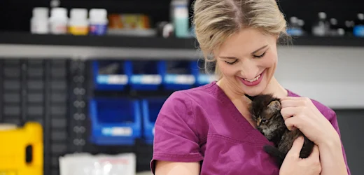 a female animal attendant with blonde hair and red lipstick in pink coloured scrubs smiles as she cradles a tiny kitten with dark fur close to her chest