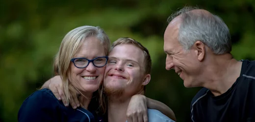 Young man with down syndrome smiles as he is hugged by his happy disability support carers