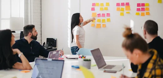 Female marketing specialist presents marketing plan and ideas using coloured post-it notes on a whiteboard, in front of the team with laptops open on conference room table