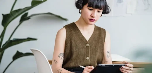 Young female graphic designer with dark hair and arm tattoos uses a stylus on a tablet to sketch out a design