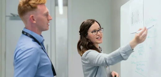A male with red hair looks on as a young female project manager colleague writes up project plans on a whiteboard at work
