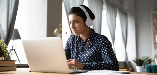 A female student focused on her online study at home, sitting in her living room wearing headphones.