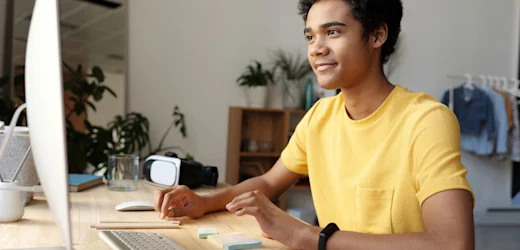A smiling young man in a yellow shirt is at his computer wondering if cyber security is a good career in Australia