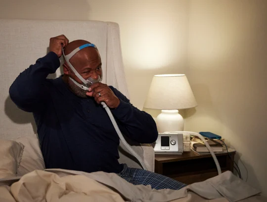 The image shows a person in a bedroom setting, adjusting a CPAP (Continuous Positive Airway Pressure) mask on their face.