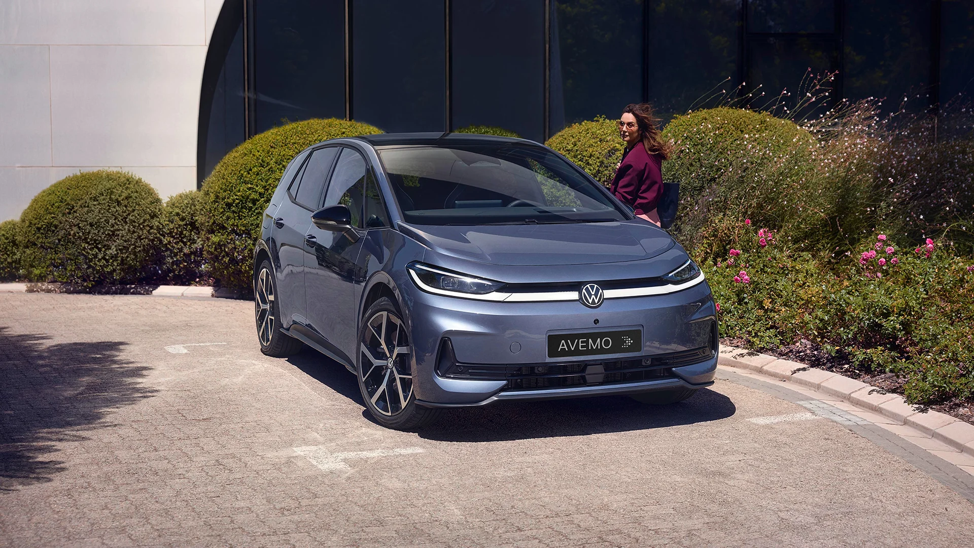 Ein blaugrauer VW ID.3 mit AVEMO-Kennzeichen parkt auf einer gepflasterten Auffahrt vor einer modernen Glasfassade. Im Hintergrund sind grün leuchtende Hecken und pinke Blumen zu sehen, während eine Frau im violetten Mantel am Auto vorbeiläuft.