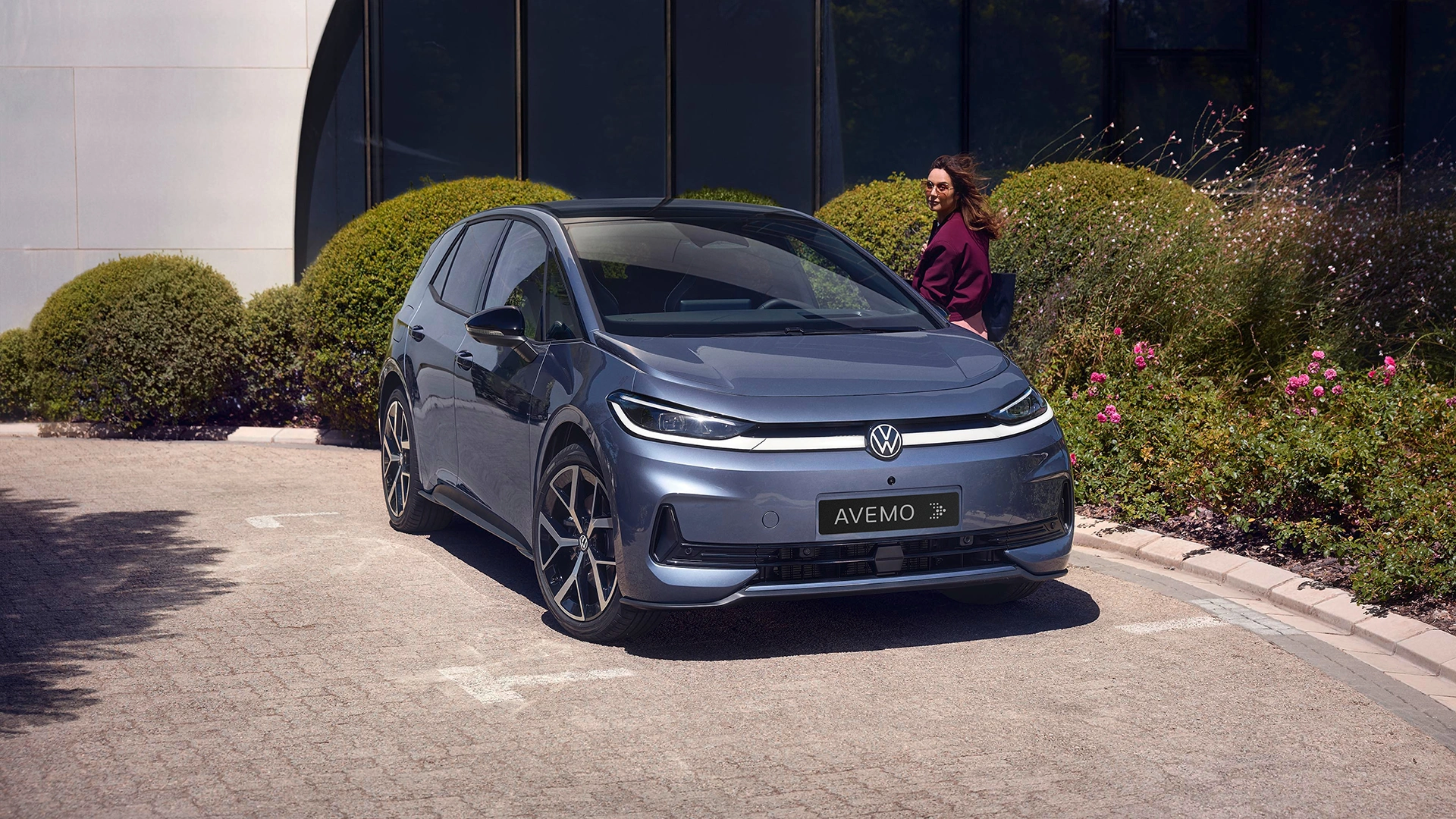 Ein blaugrauer VW ID.3 mit AVEMO-Kennzeichen parkt auf einer gepflasterten Auffahrt vor einer modernen Glasfassade. Im Hintergrund sind grün leuchtende Hecken und pinke Blumen zu sehen, während eine Frau im violetten Mantel am Auto vorbeiläuft.