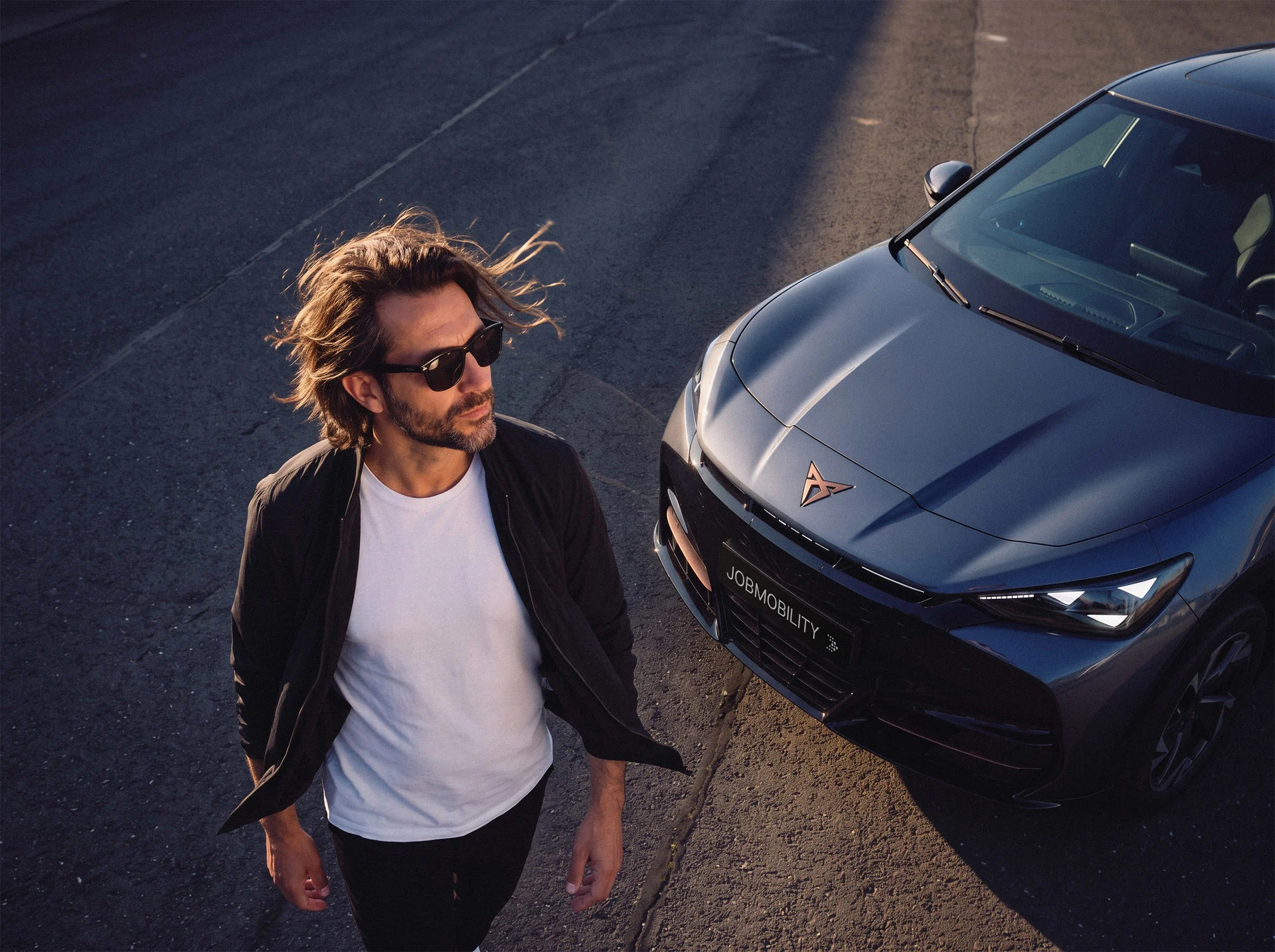 JobMobility - 1 Ein Mann mit Sonnenbrille läuft im warmen Licht neben einem sportlichen CUPRA. Das Kennzeichen zeigt JOBMOBILITY und den markanten AVEMO Pfeil.
