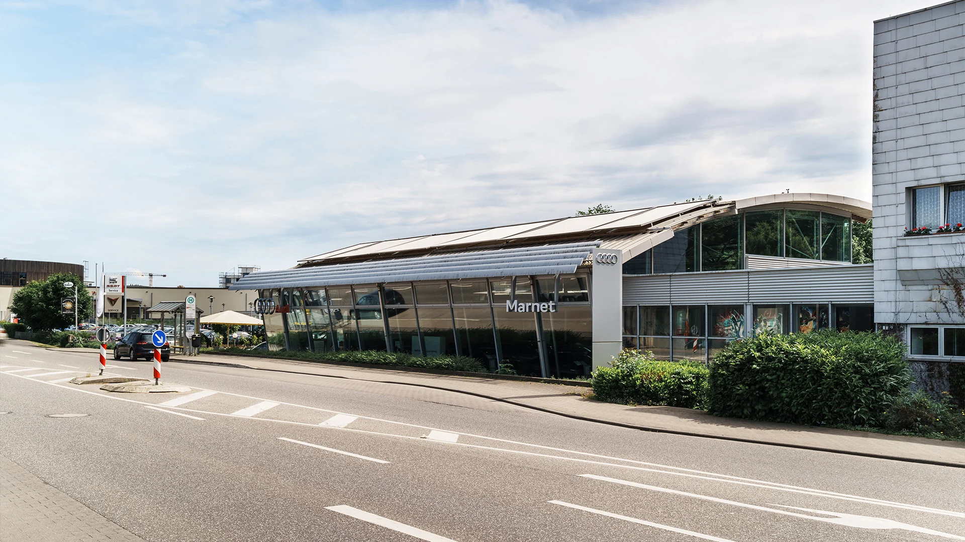 Audi Bad Nauheim Header Eine Aufnahme des Audi Autohauses in Bad Nauheim, dass das Gebäude und Audi Fahrzeuge vor dem Gebäude, sowie die Schwalheimer Straße zeigt.