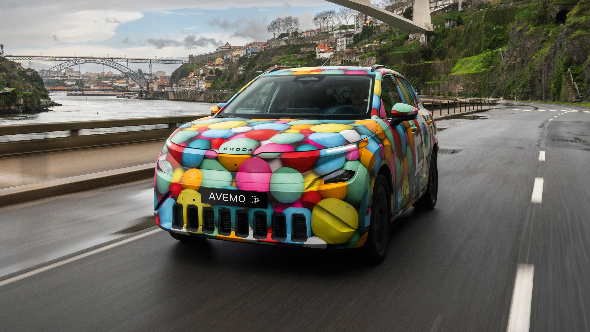 Ein Škoda Epiq mit einer auffällig bunten Folierung aus farbigen Kreisen fährt auf einer nassen Straße. Im Hintergrund ist eine Flusslandschaft mit einer markanten Brücke und Gebäuden am Hang erkennbar. Das Auto trägt ein AVEMO-Kennzeichen.