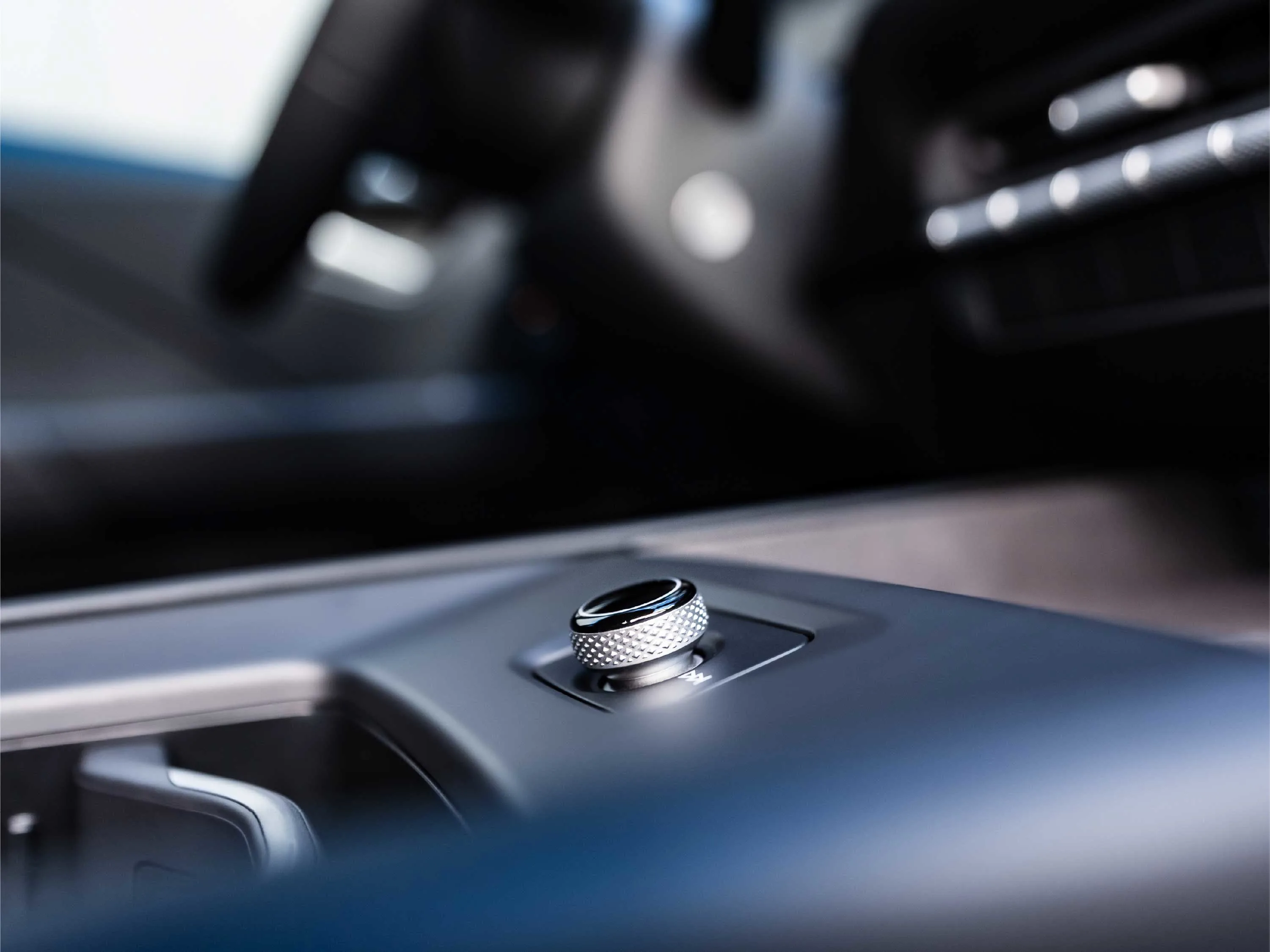 Nahaufnahme eines silbernen, geriffelten Drehreglers auf der Mittelkonsole eines VW ID.3. Das Bedienelement mit Play/Pause-Symbol ist scharf fokussiert, während das Lenkrad und Armaturenbrett im Hintergrund in Unschärfe verschwimmen.