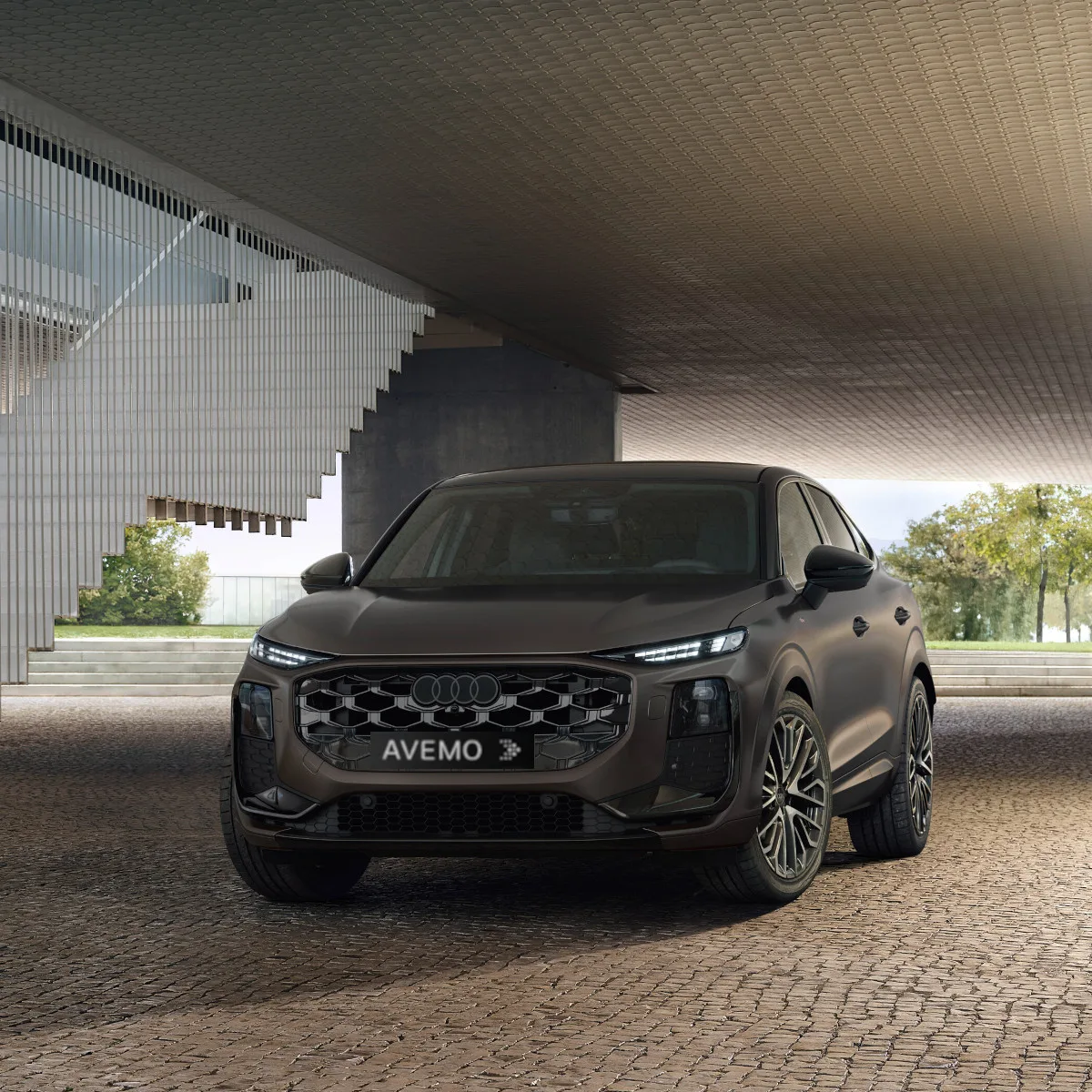Frontansicht eines dunkelbraunen Audi Q3 Sportback unter einer modernen Deckenkonstruktion auf Kopfsteinpflaster. Das Fahrzeug hat leuchtende LED-Scheinwerfer und ein AVEMO-Kennzeichen. Im Hintergrund sind eine Treppe und Grünanlagen erkennbar.
