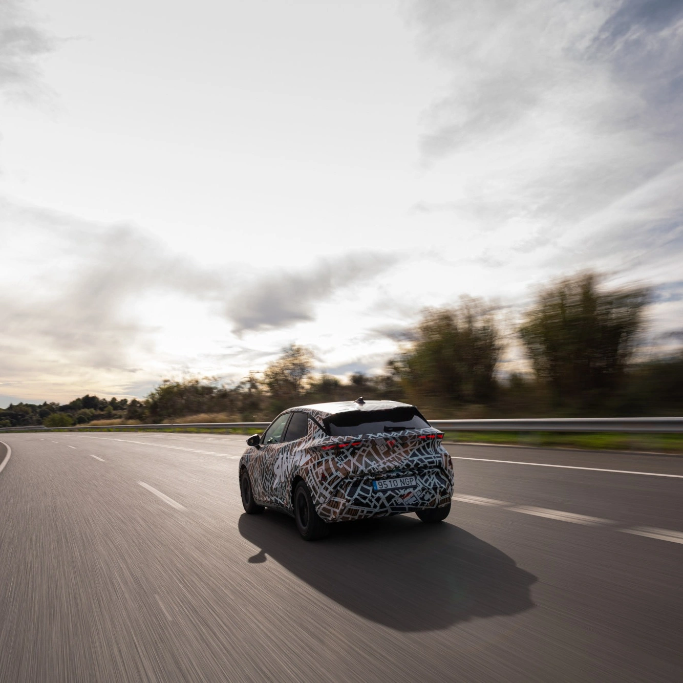 Rückansicht eines CUPRA Raval Prototyps bei einer dynamischen Testfahrt auf einer mehrspurigen Autobahn. Das kompakte Elektroauto ist mit einer auffälligen, gemusterten Tarnfolie foliert. Im Hintergrund ist ein bewölkter Himmel zu sehen.