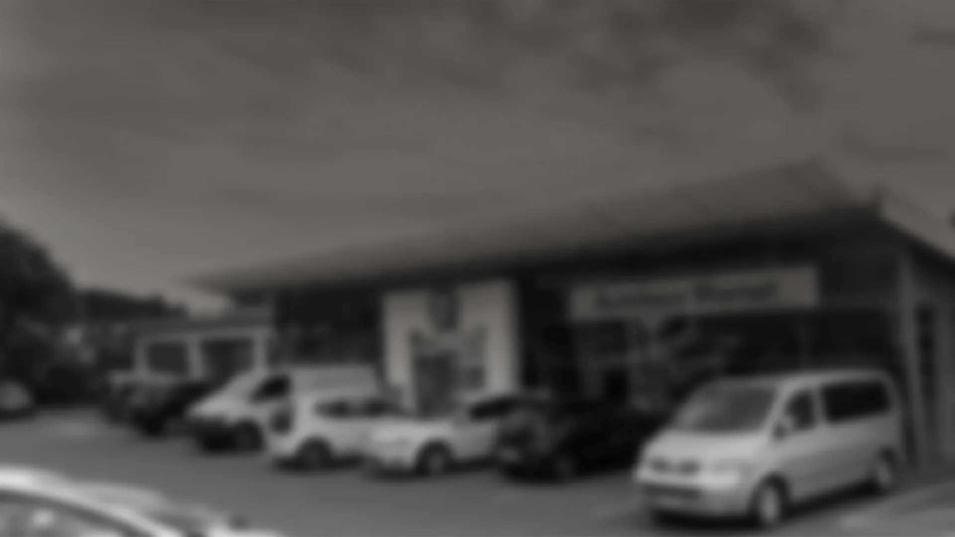 Schwarz-weiße, stark weichgezeichnete Aufnahme eines Autohauses mit davor geparkten Fahrzeugen und Transportern. Das Bild ist im Hintergrund-Stil gehalten und zeigt schemenhaft die Fassade und das Firmengelände.
