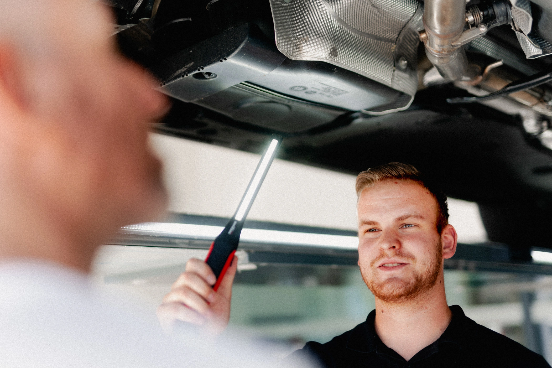 Im Bild sieht man die Schulter eines Kunden von hinten und ihm gegenüber einen Mitarbeiter des Autohauses. Sie befinden sich unter einem Auto, welches auf einer Hebebühne in der Direktannahme steht und unterhalten sich über das Fahrzeug.
