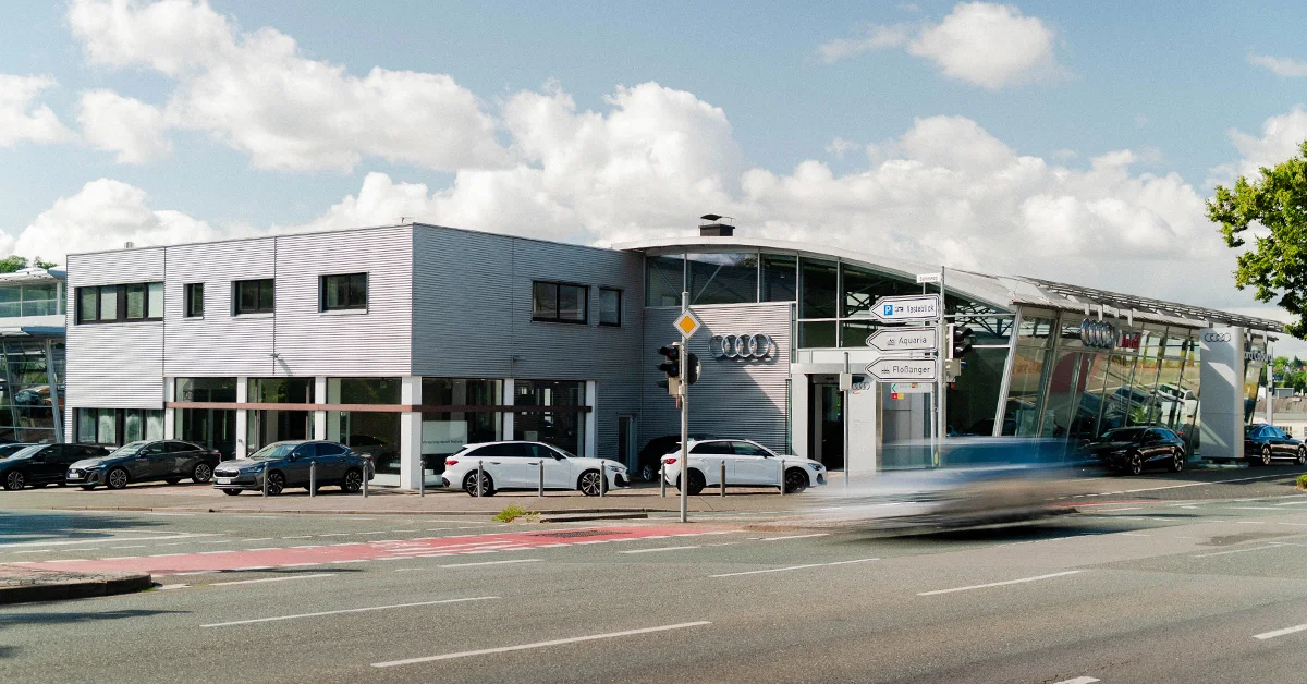 Eine Aufnahme des Audi Autohauses in Coburg, die das Gebäude und Audi Fahrzeuge vor dem Gebäude, sowie die Neustadter Straße zeigt.