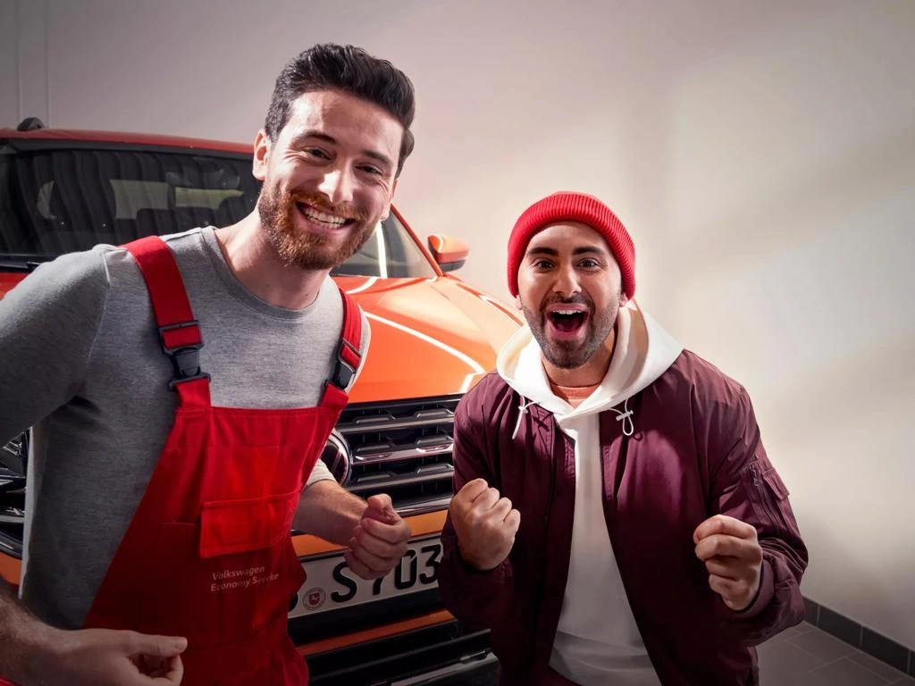economy-service Zwei lächelnde Männer feiern vor einem orangefarbenen Auto in einer Werkstatt. Einer trägt rote Arbeitskleidung, der andere einen roten Beanie und eine dunkelrote Jacke.
