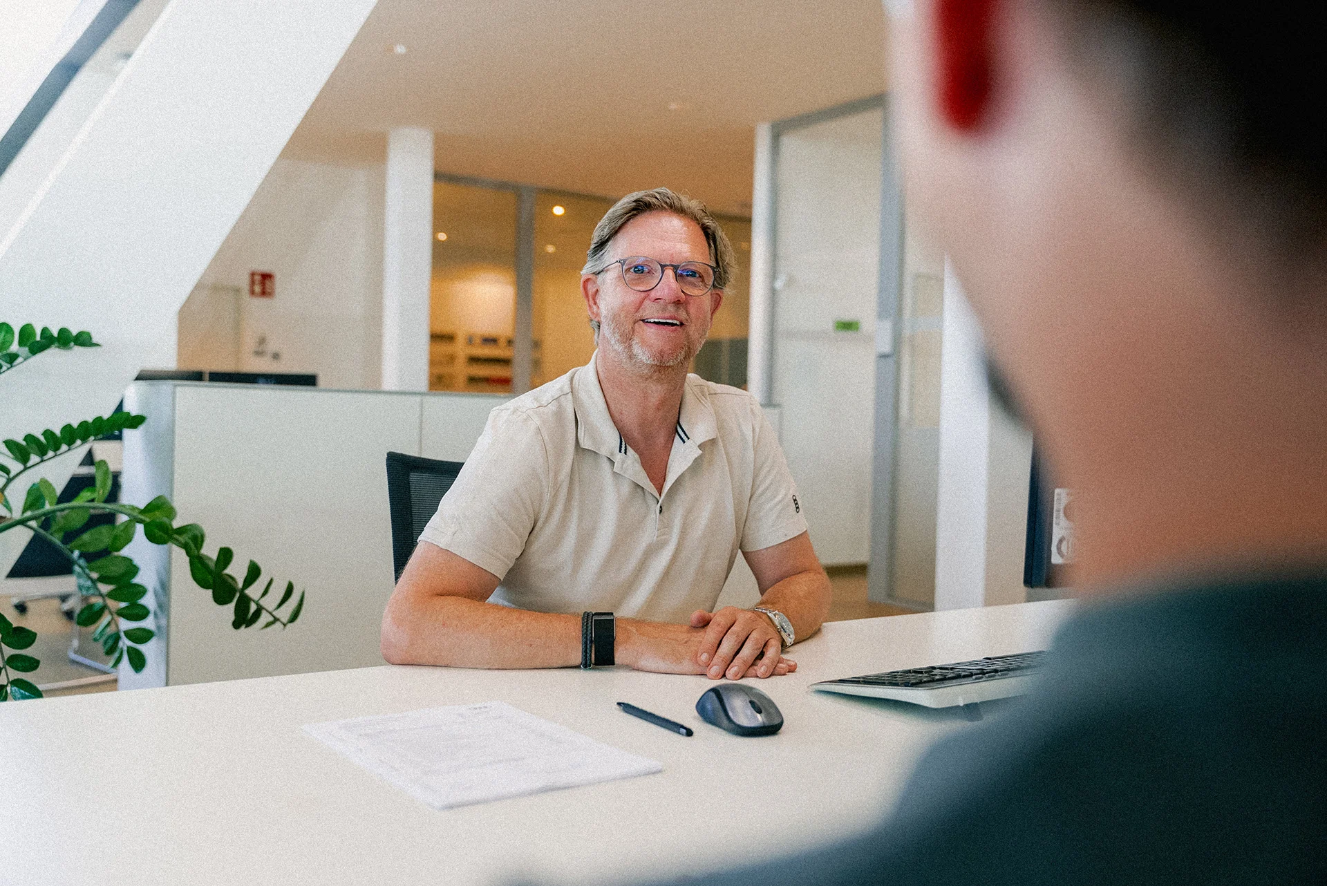 Ein Mann im beigen Poloshirt mit Brille berät einen Kunden im Showroom am weißen Tisch. Im Hintergrund sind weitere Büroräumlichkeiten mit großen Glasfenstern zu erkennen.