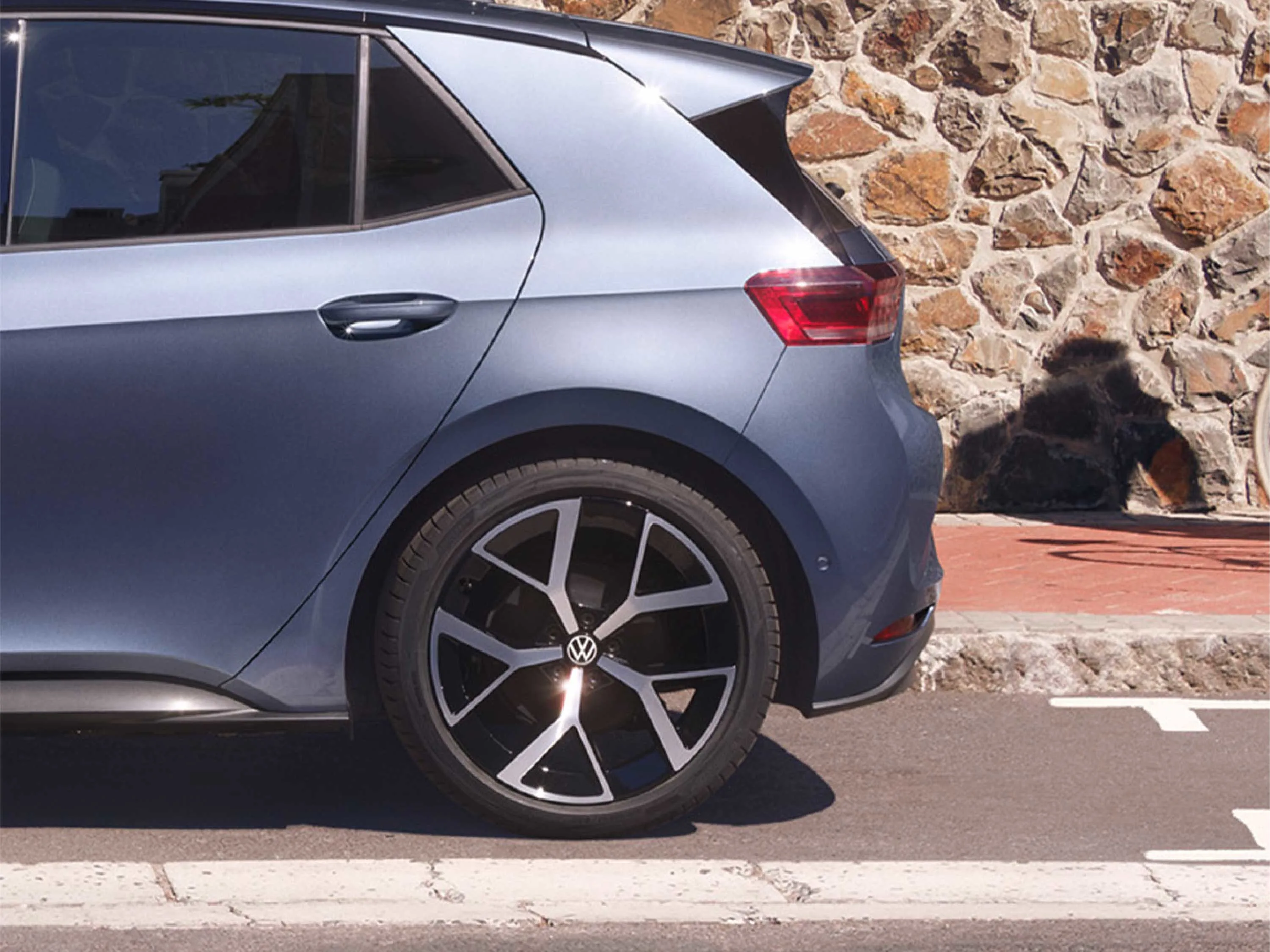 Detailaufnahme der hinteren Seitenpartie eines blaugrauen VW ID.3. Fokus auf die sportliche, schwarz-silberne Leichtmetallfelge und die markante C-Säule. Das Fahrzeug parkt vor einer rustikalen Natursteinmauer an einer Straße.
