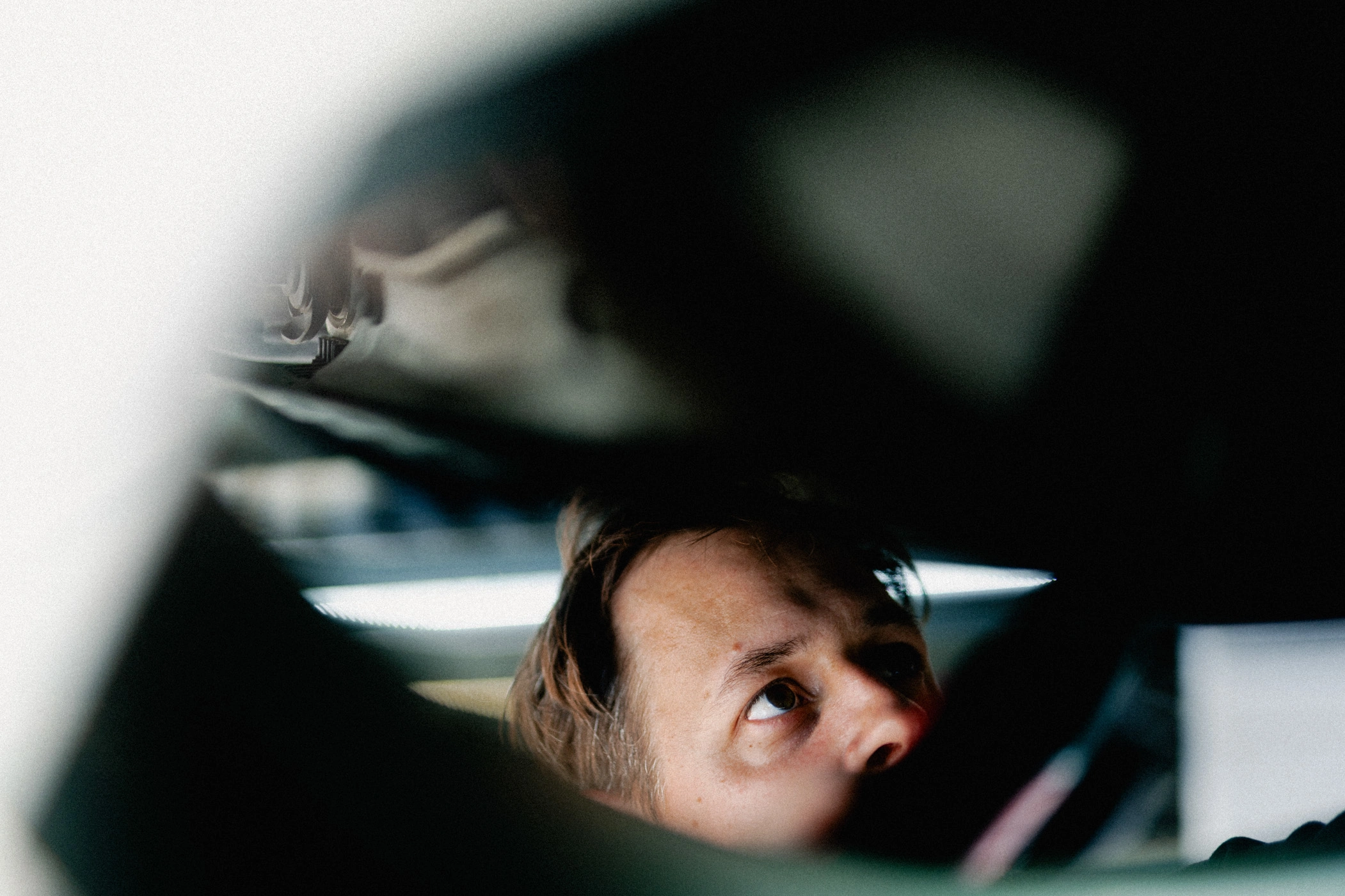 Direktannahme Hebebühne m Das Bild ist sehr abstrakt aufgenommen. Erkennen kann man nur Teile der Unterseite eines Fahrzeuges auf einer Hebebühne und einen Teil eines Gesichts eines Werkstatt-Mitarbeiters, der gerade am Auto arbeitet.