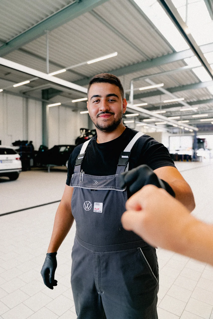 Eine Aufnahme eines Werkstatt-Mitarbeiters in dunkelblauem AVEMO Overall und schwarzen Arbeitshandschuhen, der mit dem Kameramann einen Fistbump durchführt.