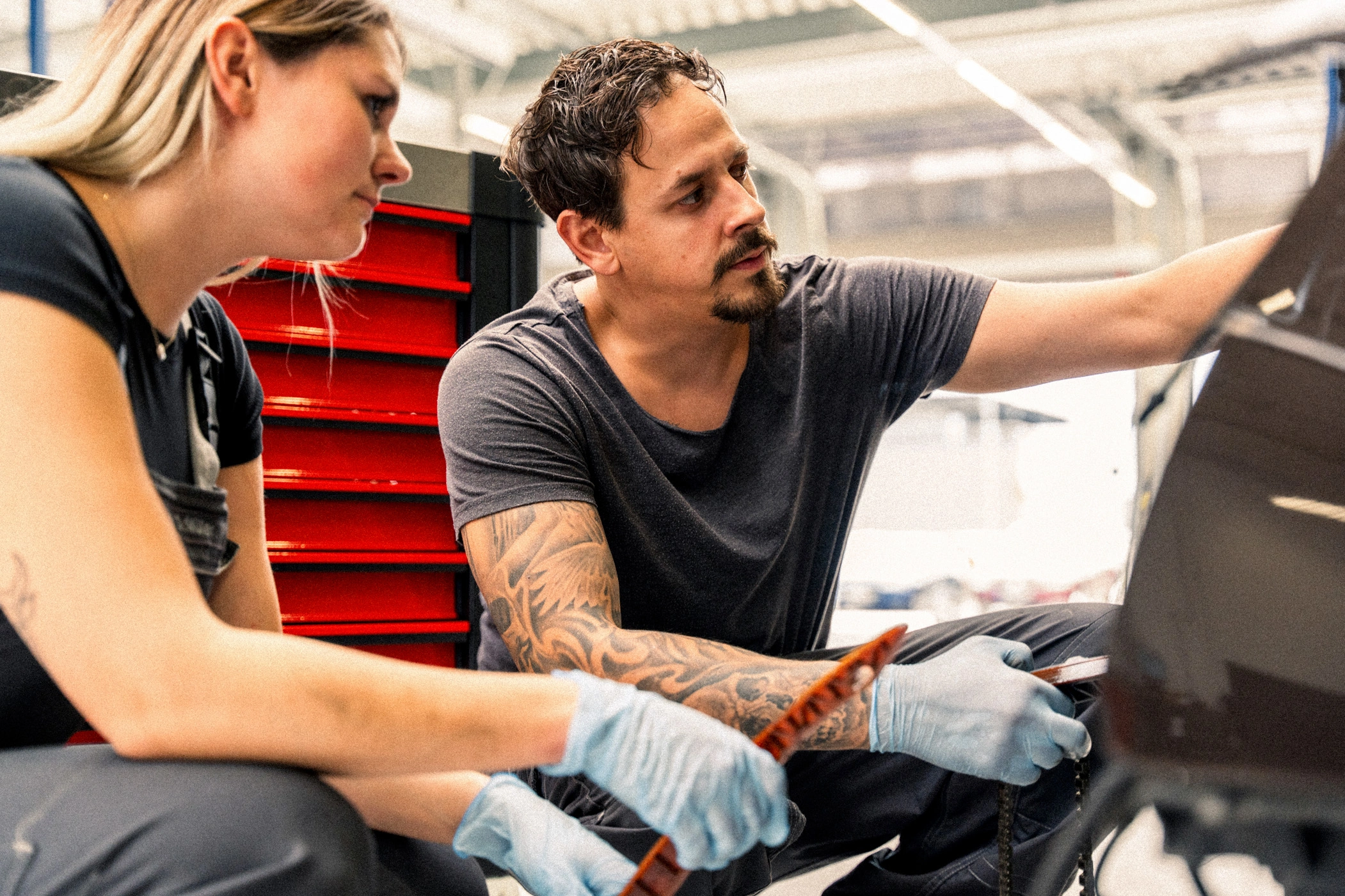 Werkstatt vorm Fahrzeug w,m Eine Frau und ein Mann knien vor einem Auto in einer Werkstatt. Beide halten Materialien in der Hand um an dem Auto zu arbeiten.