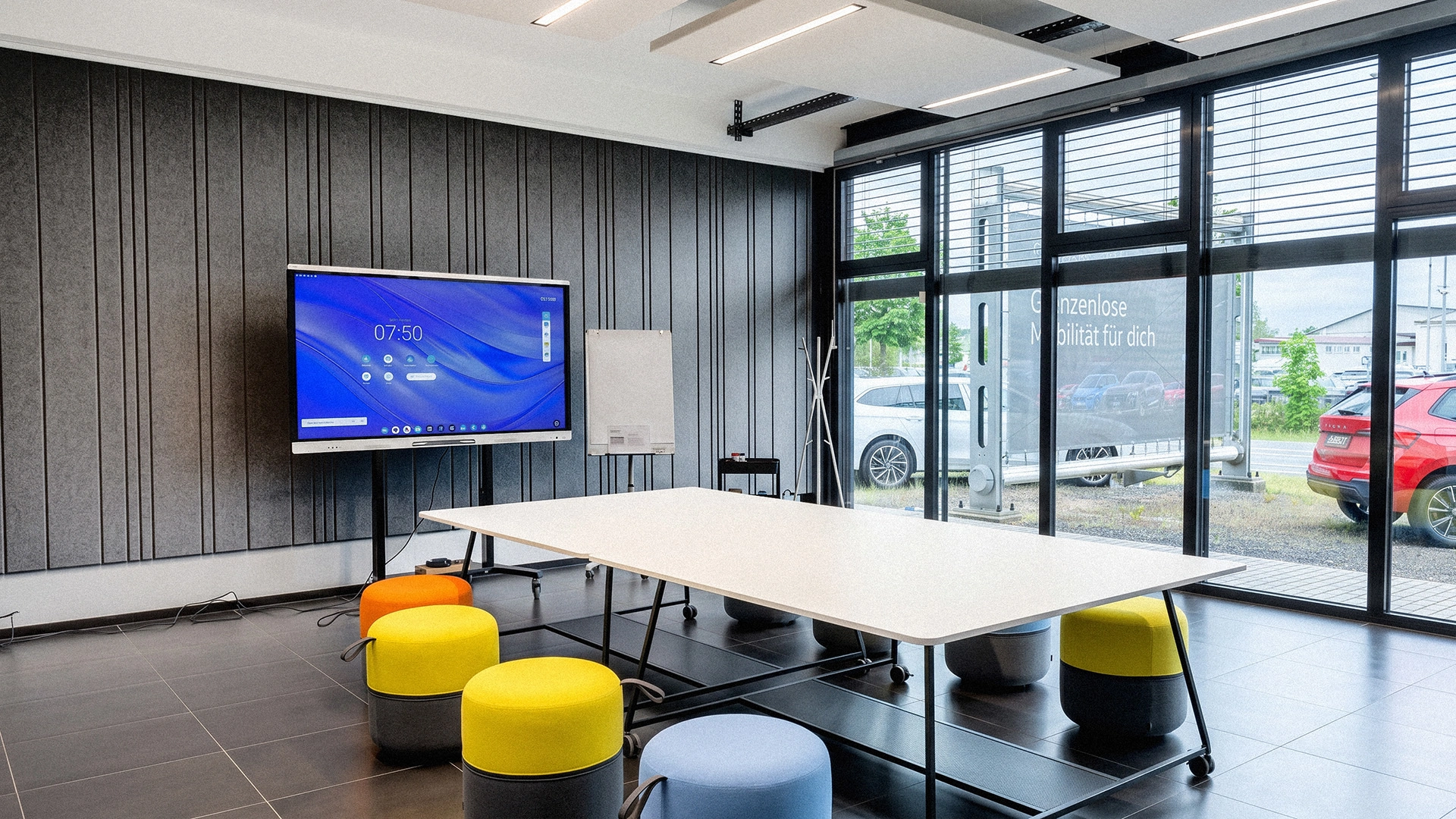 Ein heller, moderner Besprechungsraum mit einem weißen Arbeitstisch auf Rollen, bunten Sitzpoufs, einem digitalen Präsentationsbildschirm vor einer dunkelgrauen Akustikwand und einer großen Fensterfront.