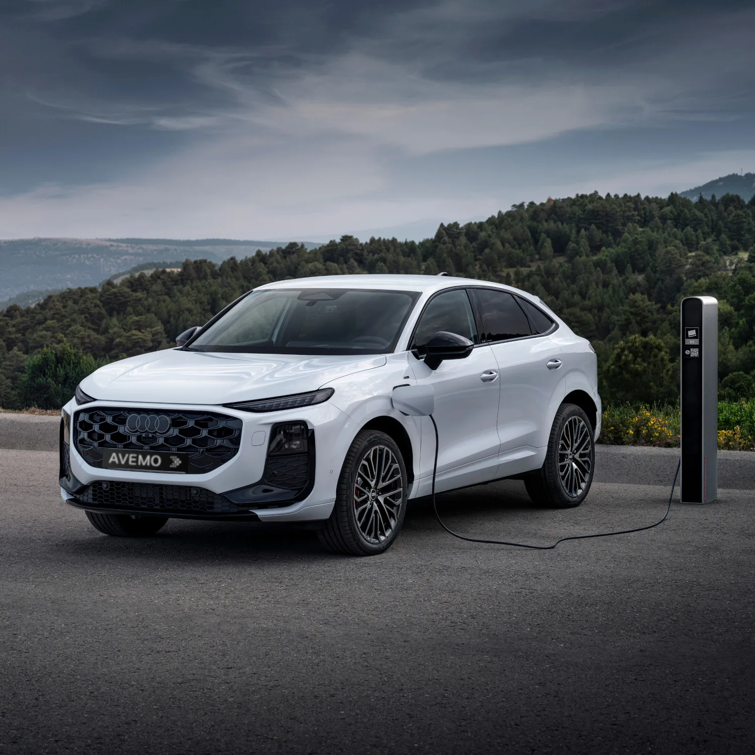 Frontansicht eines weißen Audi Q3 Sportback e-hybrid beim Ladevorgang an einer Ladesäule. Das Fahrzeug steht im Freien vor einer bewaldeten Hügellandschaft unter bewölktem Himmel und trägt ein AVEMO-Kennzeichen.