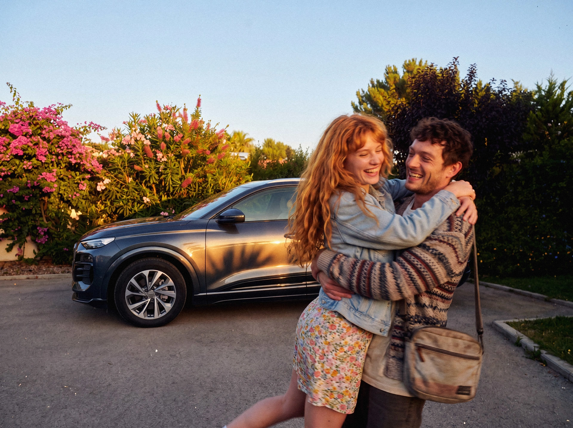 JobMobility – 12 Ein Mann im gemusterten Pullover hebt eine Frau mit roten Haaren und Jeansjacke an, sie umarmen sich lachend. Im Hintergrund steht ein dunkelgrauer Audi Q4 e-tron SUV vor grünen Büschen mit rosa Blüten.