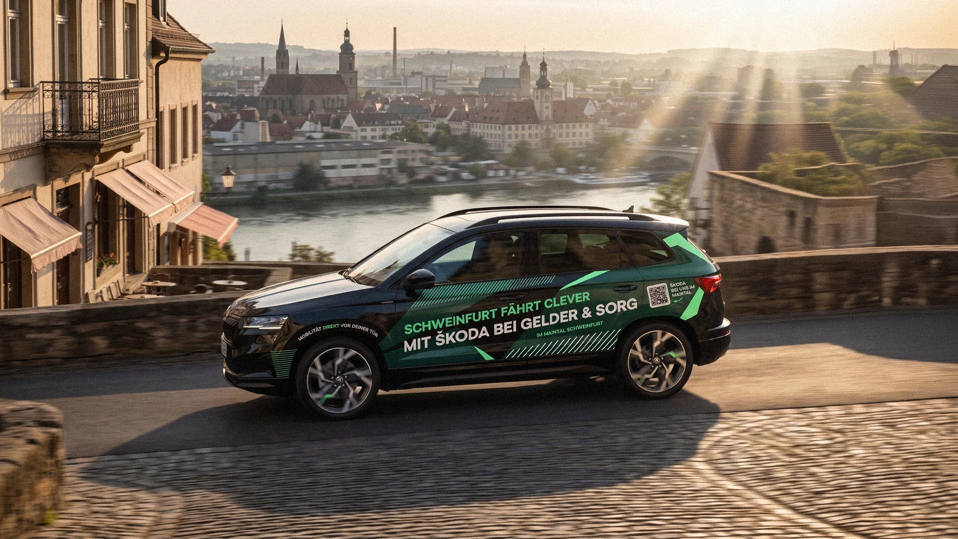 Ein schwarzer Skoda SUV fährt bei tiefstehender Abendsonne über eine gepflasterte Brücke. Im Hintergrund erstreckt sich das malerische Panorama von Schweinfurt. Die Fahrzeugbeschriftung lautet: „Schweinfurt fährt clever mit Skoda bei Gelder & Sorg“.