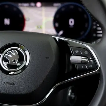 Nahaufnahme eines Skoda-Multifunktionslenkrads mit Fokus auf das Markenlogo und die silbernen Bedienelemente. Im unscharfen Hintergrund sind das digitale Cockpit mit einer Navigationskarte sowie der seitliche Infotainment-Bildschirm zu sehen.