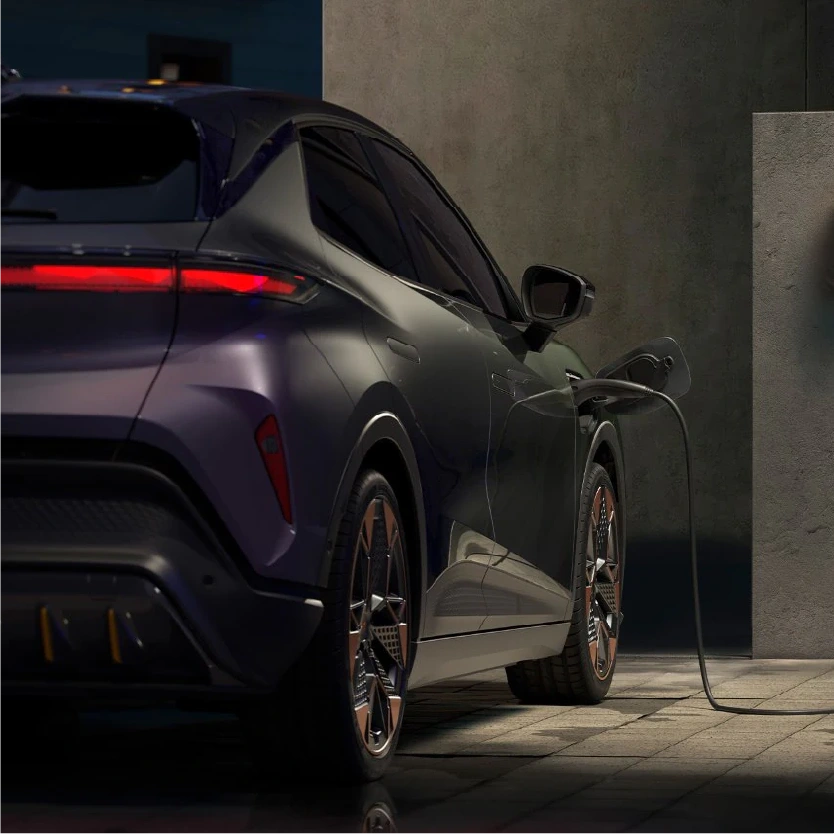 Heckansicht eines dunkelvioletten Cupra Raval, der nachts an einer Wandladestation aufgeladen wird. Das markante rote Leuchtenband am Heck ist aktiviert. Das Ladekabel ist vorne rechts am Fahrzeug angeschlossen.