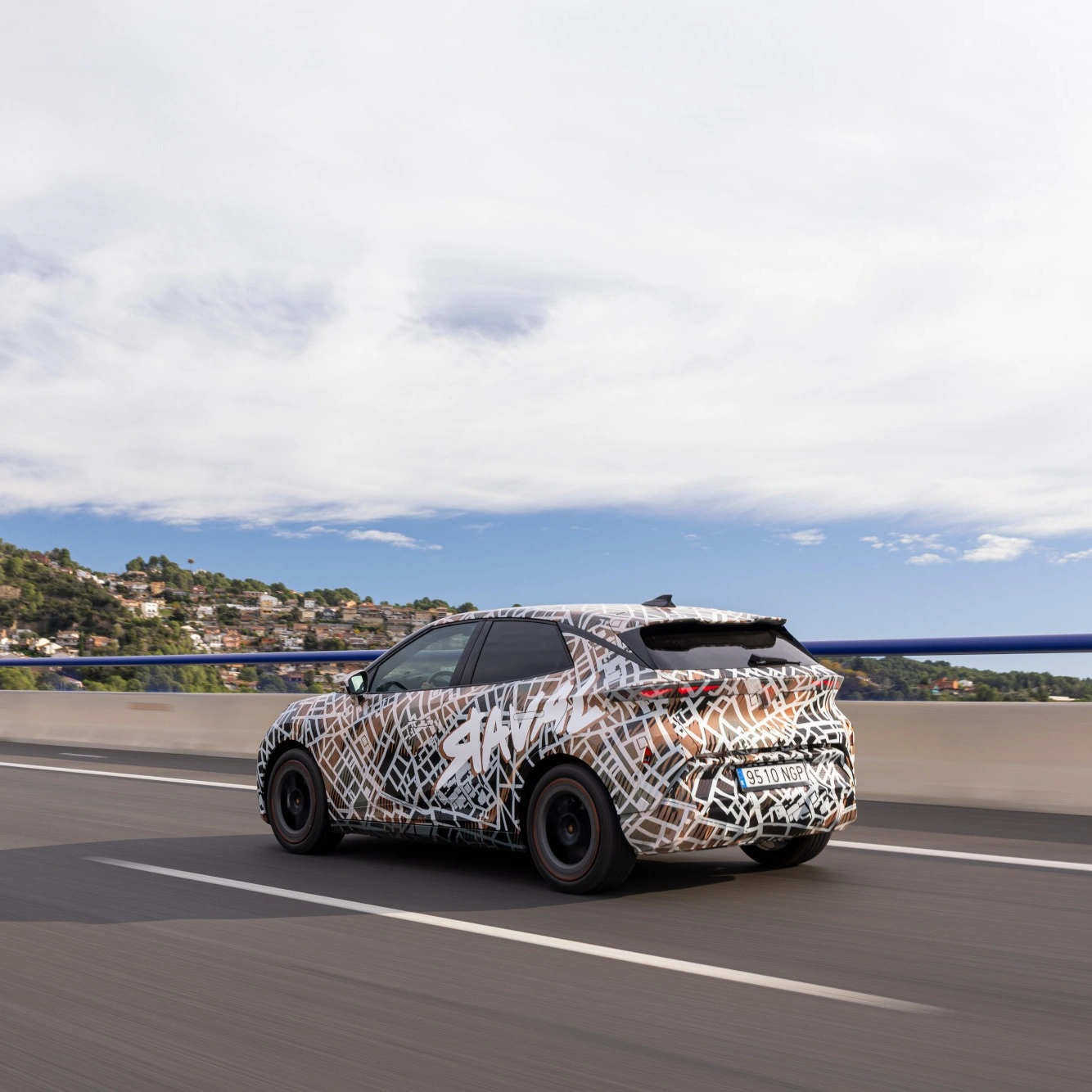 Rückansicht eines getarnten CUPRA Raval Prototyps bei der Fahrt über eine Brücke mit blauem Geländer. Die schwarz-weiß-braune Tarnfolie verbirgt das Design des Elektroautos. Im Hintergrund ist eine bewaldete Hügellandschaft mit Häusern zu sehen.