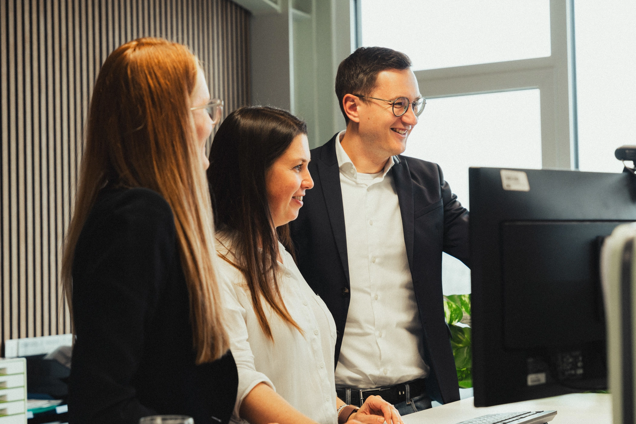 Drei Büro-Mitarbeitende stehen an einem höhenverstellbaren Schreibtisch, um gemeinsam ein Online-Meeting abzuhalten. Außerdem sieht man eine moderne Büroeinrichtung mit Pflanzen und Holzleisten an den Wänden.