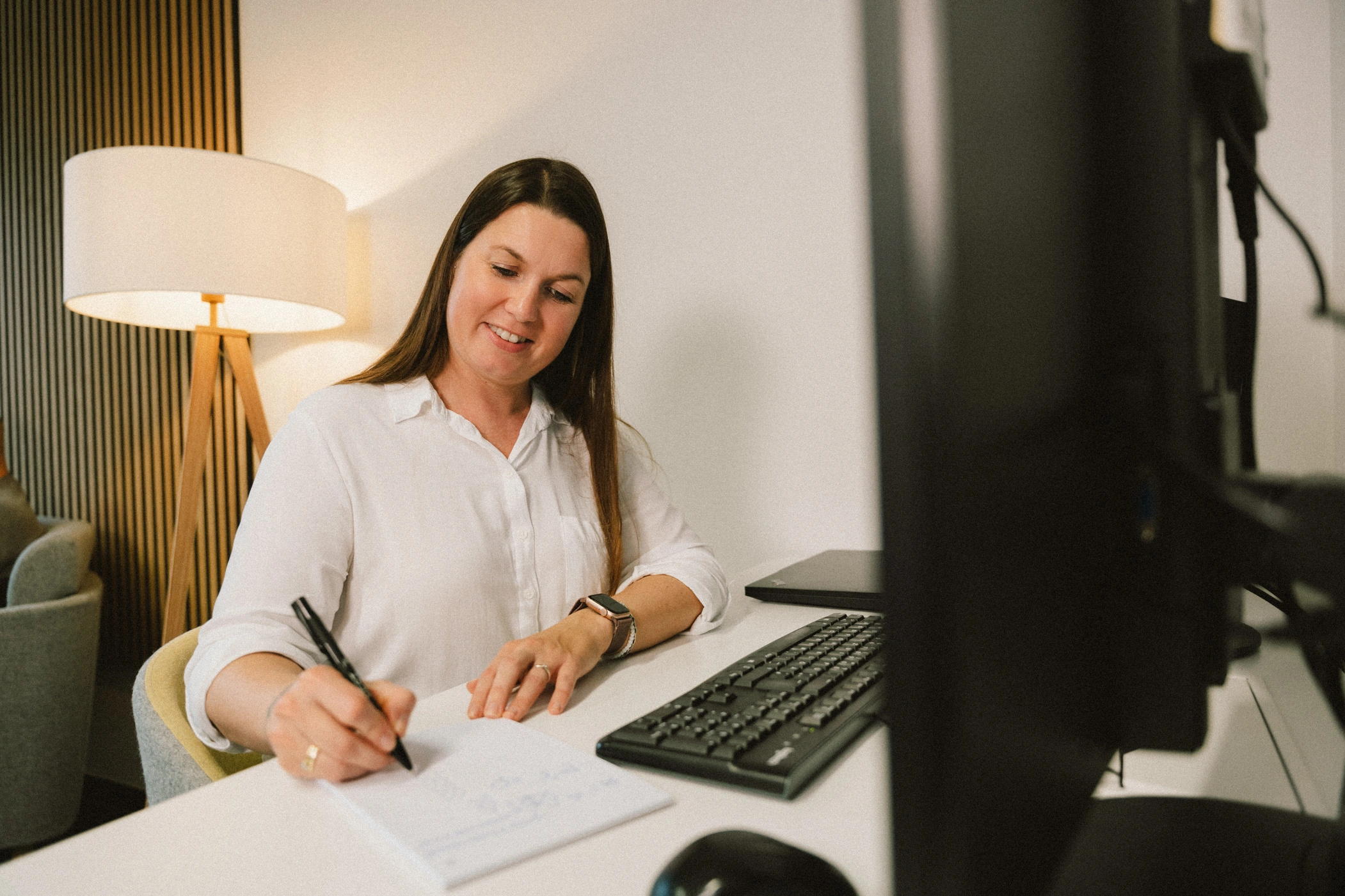 Büro Block aufschreiben (2) Eine Frau in weißer Bluse sitzt an einem Schreibtisch und notiert etwas auf einem Notizblock. Der Schreibtisch ist eingerichtet wie ein typischer Arbeitsplatz mit Monitoren und weiterem PC-Equipment. Im Hintergrund steht eine Lampe.