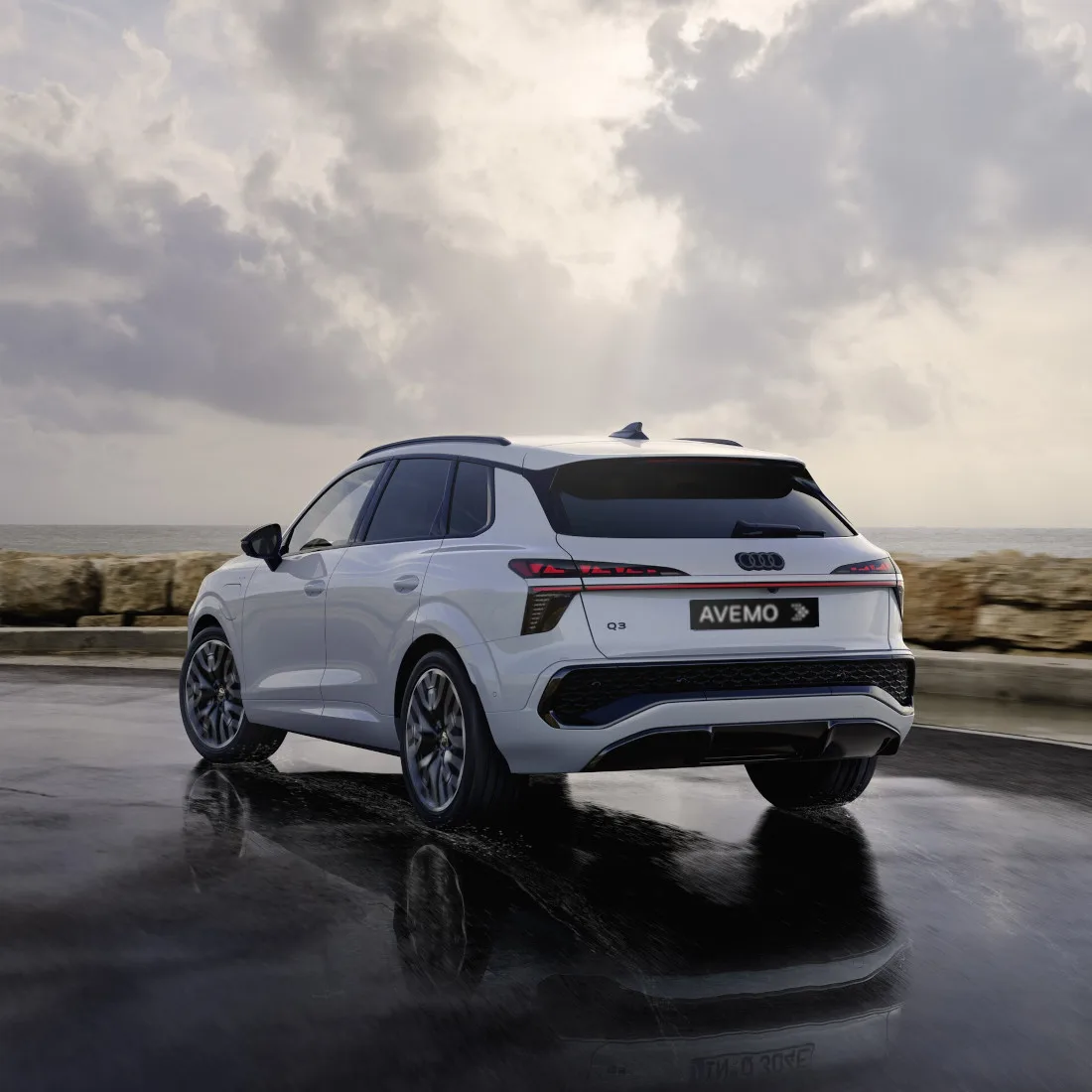 Heckansicht eines weißen Audi Q3 SUV auf einer nassen Straße am Meer unter einem dramatisch bewölkten Himmel. Das Fahrzeug spiegelt sich auf dem feuchten Asphalt und trägt ein AVEMO-Kennzeichen sowie markante LED-Rückleuchten.