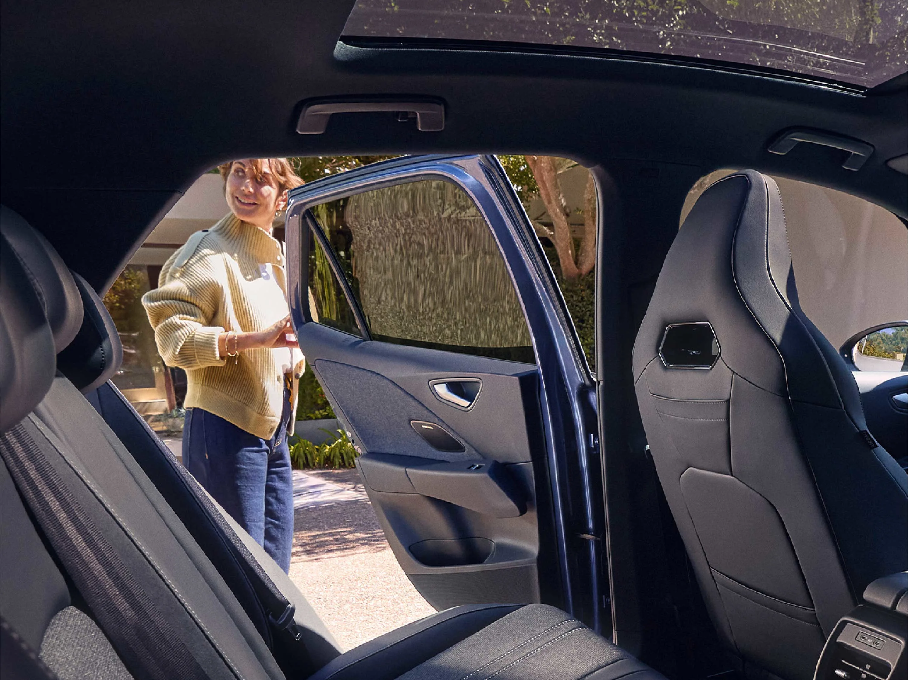 Blick durch den Fond eines VW ID.3 bei geöffneter Fondtür. Eine lächelnde Frau steht draußen neben dem Fahrzeug. Der Innenraum zeigt dunkle Sportsitze mit integrierten Kopfstützen und das Panorama-Glasdach lässt helles Tageslicht herein.