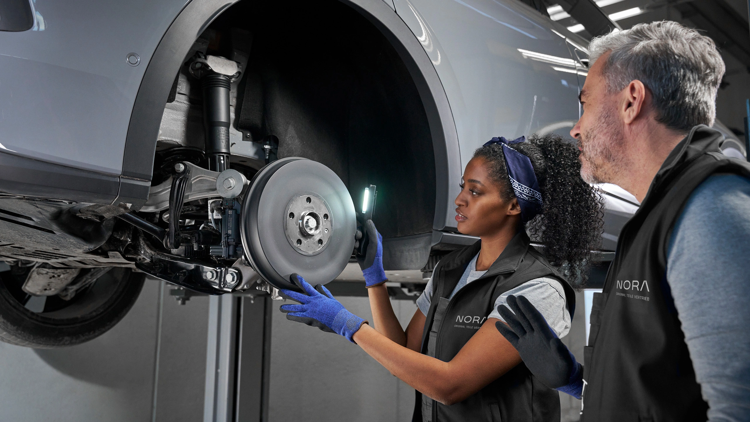 Zwei Fachkräfte in einer Werkstatt inspizieren die Bremsanlage eines angehobenen Fahrzeugs. Beide tragen schwarze Arbeitswesten mit dem „NORA“-Logo. Eine Mechanikerin leuchtet mit einer Lampe in den Radkasten, während ein Kollege die Arbeit begutachtet.