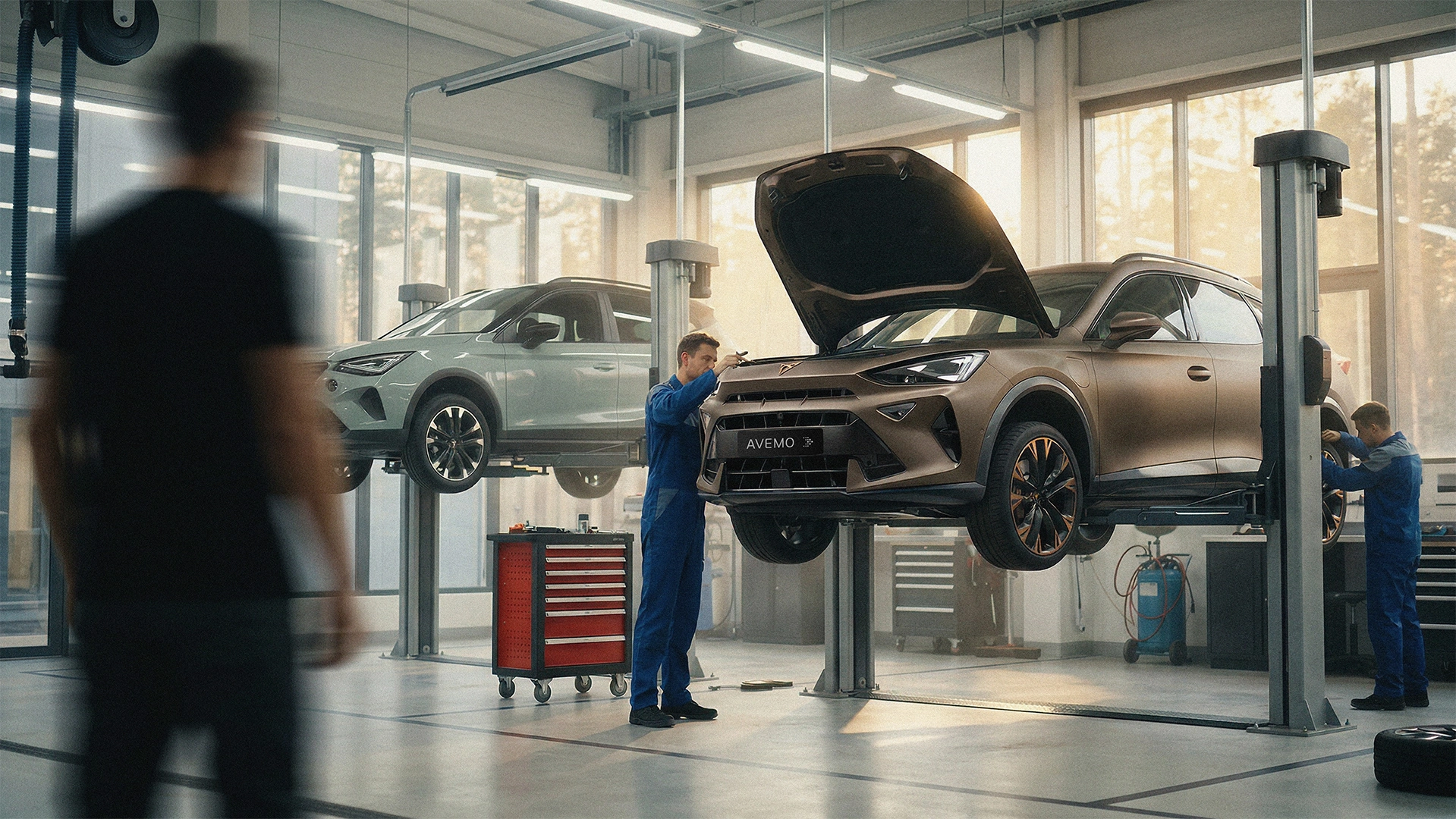 A23-0065 website header Blick in eine moderne, lichtdurchflutete Werkstatt. Zwei Fahrzeuge, darunter ein bronzefarbener Cupra mit geöffneter Motorhaube, befinden sich auf Hebebühnen. Mechaniker in blauer Arbeitskleidung führen Servicearbeiten an den Autos durch.