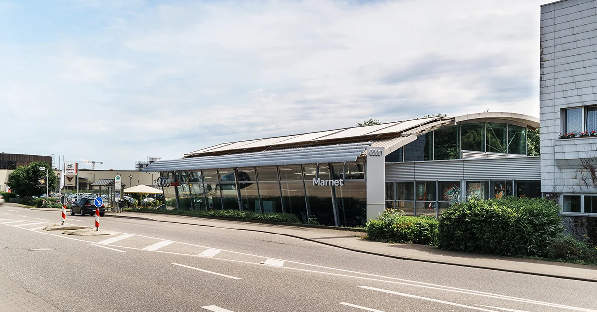 Eine Aufnahme des Audi Autohauses in Bad Nauheim, die das Gebäude und Audi Fahrzeuge vor dem Gebäude, sowie die Schwalheimer Straße zeigt.