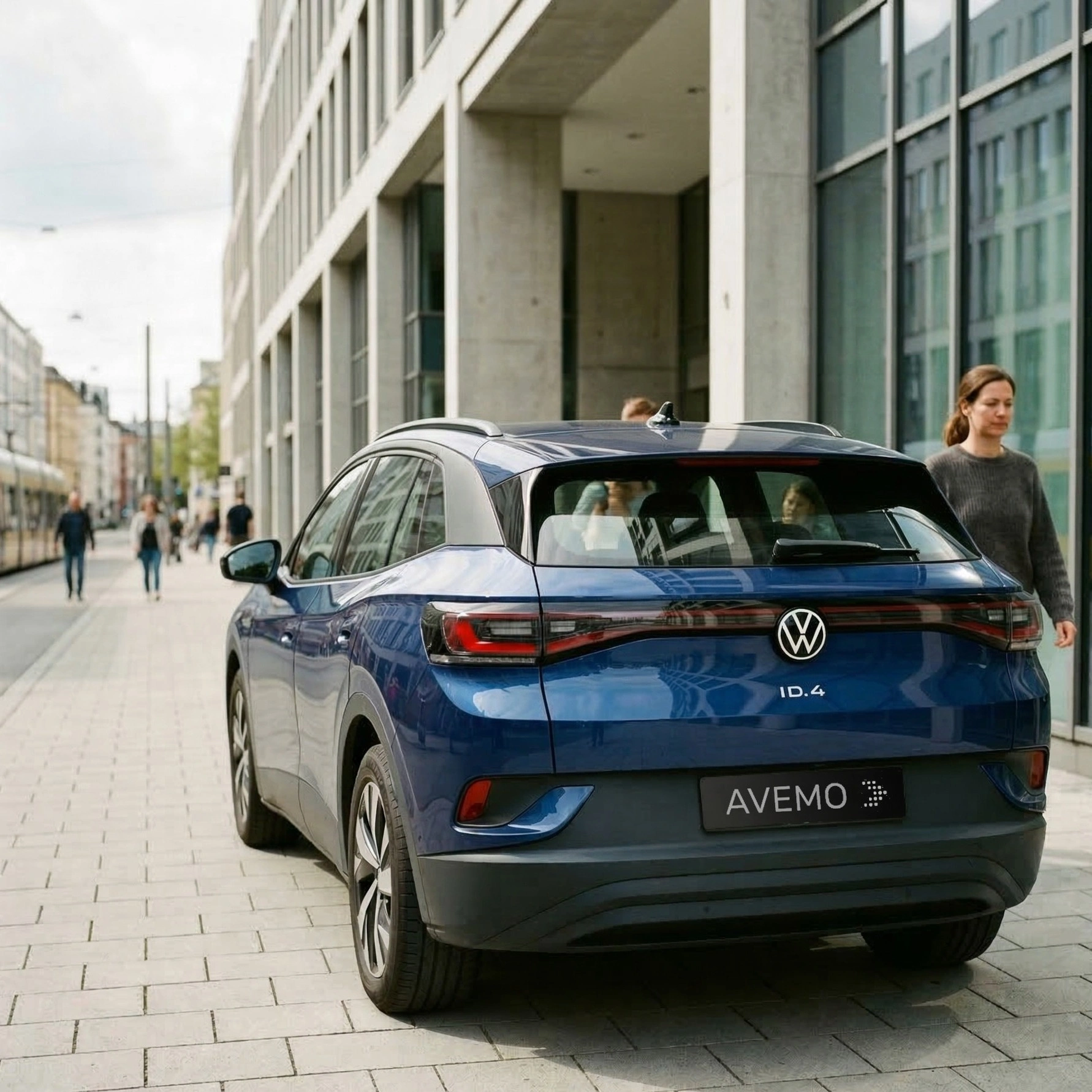 Rückansicht eines blauen Volkswagen ID.4 mit AVEMO-Kennzeichen, geparkt auf einem gepflasterten Gehweg vor einem modernen Bürogebäude mit Glasfassade. Passanten und eine angedeutete Straßenbahn im Hintergrund unterstreichen die urbane Szene.