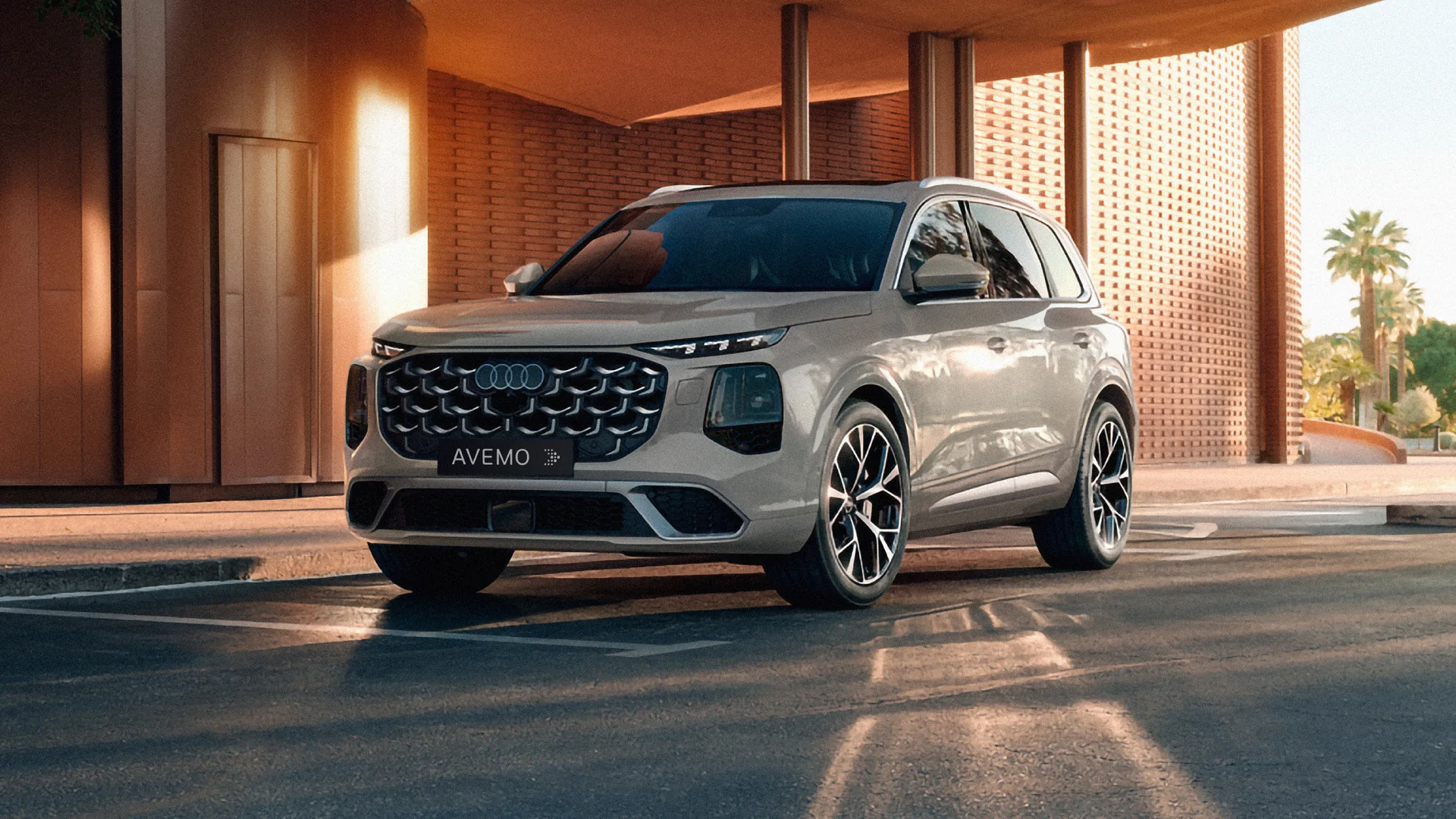 Ein beigefarbener Audi SUV parkt vor einem markanten Backsteingebäude. Das Fahrzeug ist von schräg vorne zu sehen, zeigt den großen, dunklen Kühlergrill mit den Audi-Ringen und auffällige Alufelgen, während im Hintergrund Palmen erkennbar sind.