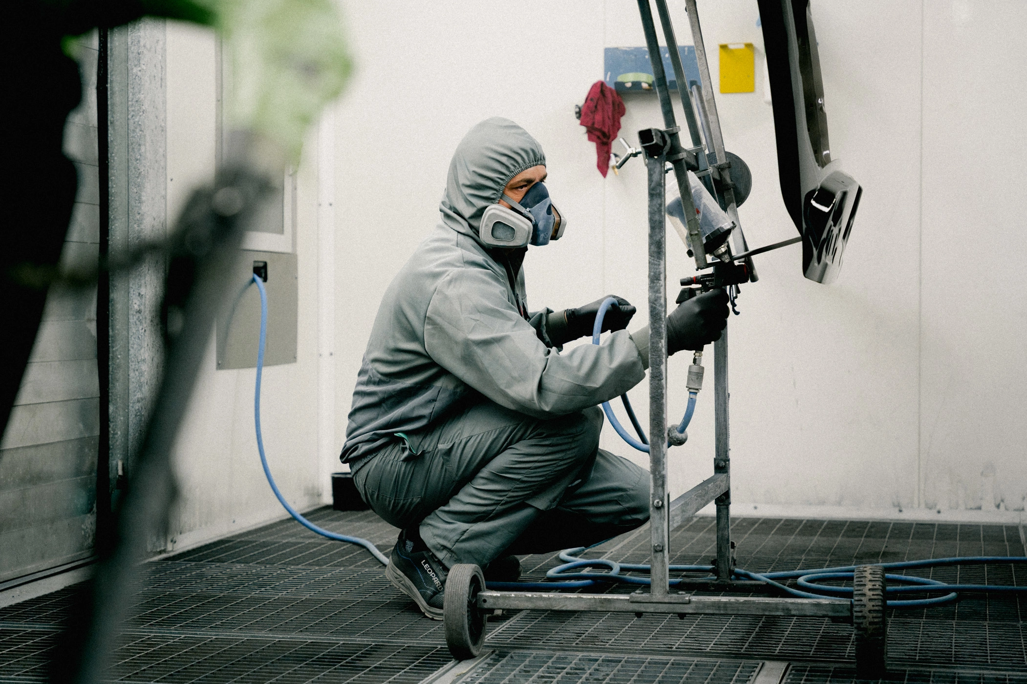 Lackierkabine m-2 Man sieht einen Mitarbeiter in Schutzanzug und mit Schutzmaske, der vor einem Karosserie-Teil in der Lackierkabine hockt. In seiner rechten Hand hält er eine Sprüh-Pistole mit Klarlack, den er auf das Bauteil aufträgt.